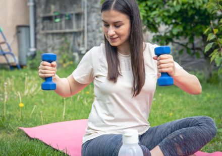 Sportive young woman stretching with dumbbells, doing fitness exercises in green park.